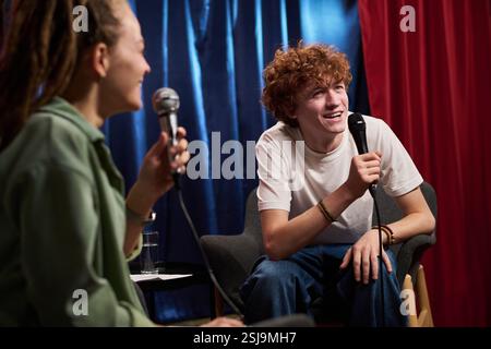 Porträt eines jungen Mannes mit lockigen Haaren, der während des Interviews neben der Person mit geflochtenem Haar auf einem Sessel sitzt und ein Mikrofon hält Stockfoto
