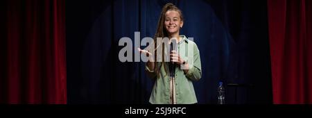 Frau, die auf der Bühne steht und lächelt, während sie das Mikrofon in der Hand hält und einen freudigen Moment während der Vorstellung vor dunkler Kulisse teilt Stockfoto