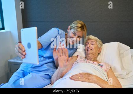 Eine Krankenschwester, die eine ältere Frau in einer Seniorenrehabilitationseinrichtung unterstützt und mit einem Tablet Videogespräche führt. Der Moment fängt Aufmerksamkeit ein, Stockfoto