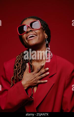Lachende afroamerikanische Frau mit stilvoller roter Sonnenbrille und großen Creolen vor dunkelrotem Hintergrund, die einen freudigen Moment feiert und genießt Stockfoto