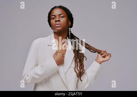Porträt einer selbstbewussten Afroamerikanerin, die das Haar anpasst und gleichzeitig einen stylischen weißen Blazer im Studio trägt. Lange Zöpfe und elegante Ohrringe sorgen für ein einzigartiges Flair Stockfoto
