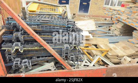 Ein rostroter Behälter enthält eine gemischte Auswahl von Baumaterialien, überwiegend Stapel von schwarzen Gittern, Holzdielen und Metallgerüsten Stockfoto