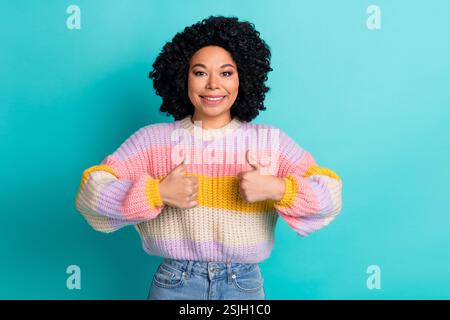 Glückliche junge Frau mit lockigem Haar, die einen farbenfrohen Pullover trägt, der die Daumen hoch vor einem blaugrünen Hintergrund zeigt Stockfoto