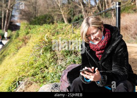 Anderlecht, Region Brüssel-Hauptstadt - Belgien : 12 25 2020 attraktive dreißigjährige Frau, die in einem sonnigen Park das Smartphone überprüft. Modellversion Stockfoto