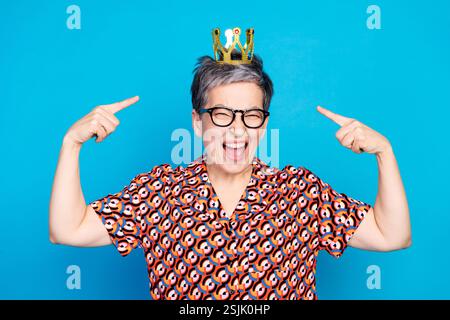 Aufgeregte Seniorin, die in einem bunten Outfit auf ihre Krone zeigt und vor einem hellblauen Hintergrund lächelt Stockfoto