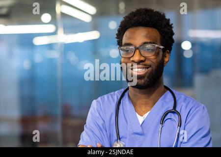 Nahporträt eines jungen erfahrenen Arztes. Ein Mann mit einem Stethoskop lächelt und schaut zur Seite, gekleidet in einem blauen medizinischen Kleid. Der Arzt arbeitet im Büro einer modernen Klinik. Stockfoto