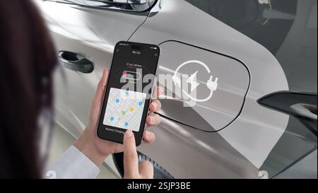 Frau überprüft ihre EV-App auf einem Smartphone und zeigt eine Warnung bei niedrigem Batteriestand und nahe gelegene Ladestationen an, während ein Elektrofahrzeug im Hinterland geparkt ist Stockfoto
