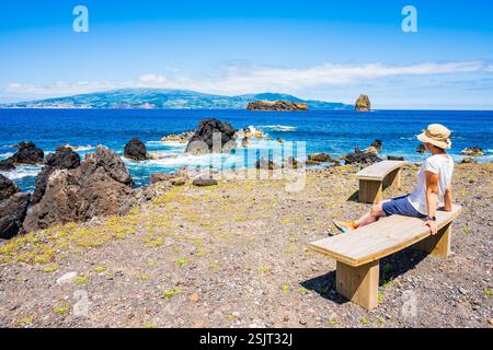Junge Touristin mit Hut auf der Bank und Blick auf das Meer an der Küste der Insel Pico, Azoren, Portugal Stockfoto