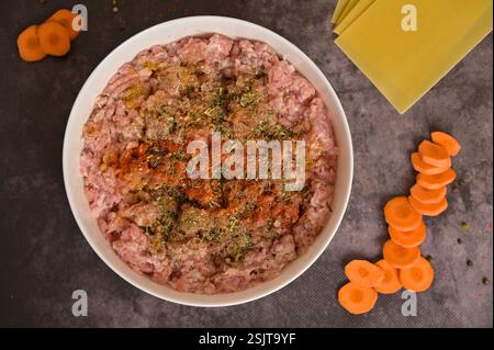 Roh gemahlenes Hackfleisch von oben in einer weißen Schüssel mit Gewürzen. Lasagne Nudelblätter, Karotten, Pfeffer. Schwarzer Hintergrund. Hausgemachte Lasagne-Küche Stockfoto