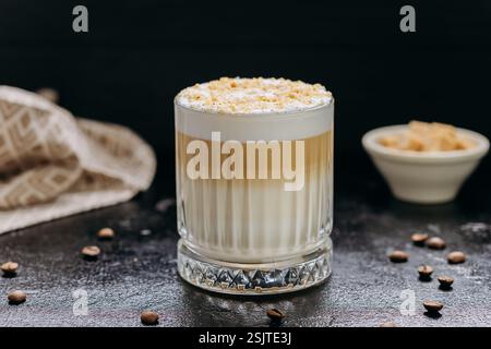 Lagenlatte mit aufgeschäumter Milch und zerstoßenen Nüssen in Kristallglas mit Kaffeebohnen auf dunklem Tisch Stockfoto