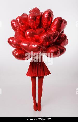 Stilvolle Frau in Rot, die einen Haufen herzförmiger Ballons hält und ihr Gesicht versteckt. Romantisches, festliches und Valentinstag inspiriertes Konzept. Stockfoto