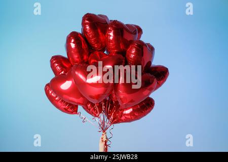 Eine Hand hält einen Haufen roter Herzballons vor einem leuchtend blauen Himmel, die Liebe, Feier und Freude symbolisieren. Stockfoto