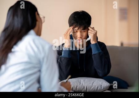 Ein gestresster, weinender junger asiatischer Mann spricht mit seinem Psychologen in der Klinik und erlebt einen psychischen Zusammenbruch während einer Untersuchung. Geistig Stockfoto