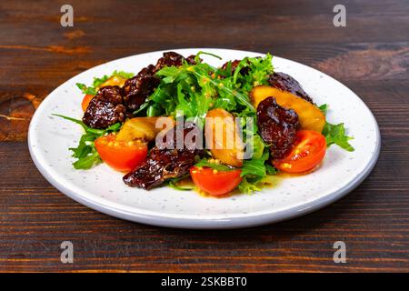 Ein lebhafter Salat mit einer Mischung aus Rucola, glasierten Früchten und Kirschtomaten, ordentlich auf einem weißen Teller angeordnet. Das Gericht wird auf einem rustikalen Holz serviert Stockfoto