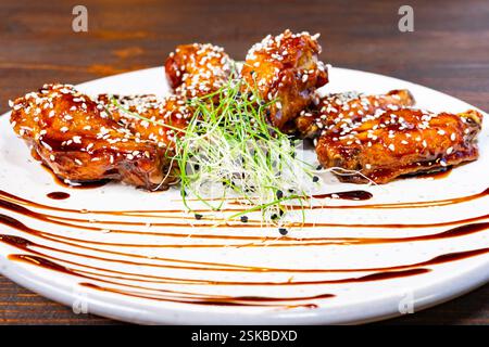 Ein Teller zeigt saftige Hühnerflügel, glasiert mit einer reichhaltigen Sauce und garniert mit Sesamsamen und frischem Gemüse. Diese kulinarische Köstlichkeit ist perfekt für Sie Stockfoto