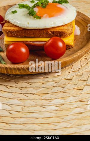 Auf einem Holzteller wird ein köstliches Frühstückssandwich mit geröstetem Brot, geschmolzenem Käse und einem sonnigen Ei serviert. Frische Tomaten werden zubereitet Stockfoto