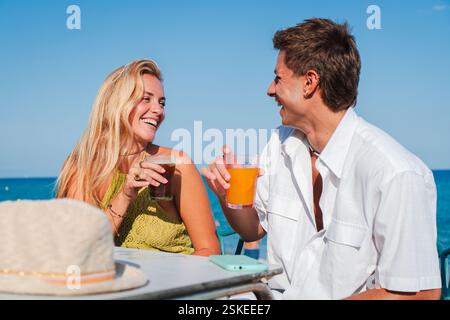 Fröhliches Paar genießt erfrischende Getränke, während es sich an Einem hellen sonnigen Tag verspielt am Beachside Table antost, während Lachen und Lächeln ihre Gesichter und Herzen voller Glück erhellen. Hochwertige Fotos Stockfoto