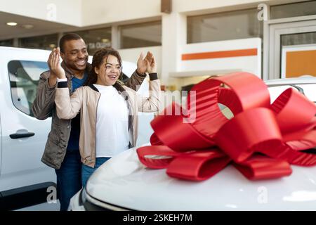 Eigenes Auto. Afrikanischer amrerikanischer Ehemann bedeckt die Augen seiner Frau und zeigt ihr neues Auto im Händlerladen. Leerer Bereich Stockfoto