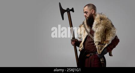 Mittelalterlicher Wikinger, gepanzert, mit massiver Axt, mit harter Entschlossenheit vor grauem Atelierhintergrund. Stockfoto