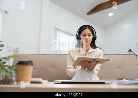 Junge Frau, die Kopfhörer trägt, während sie Notizen auf einer Couch liest, in ihre Lernsitzung eingetaucht. Stockfoto