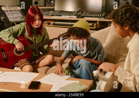 Männliches Mitglied der Teenager-Band, das Liedtexte liest, während seine Gitarristinnen sich Texte anschauen und versuchen, Melodien zu entwickeln Stockfoto