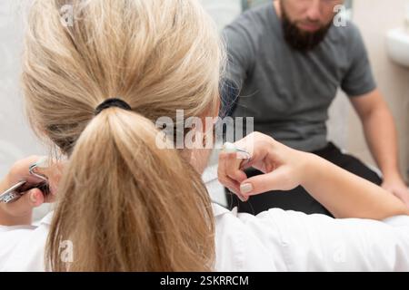Blick von hinten auf eine hellhaarige Frau mit einem Pferdeschwanz, der Zahnwerkzeuge hält. Er steht vor einem Mann mit Bart, der in einem Stuhl sitzt Stockfoto