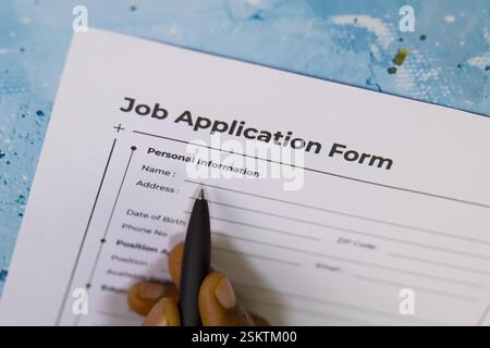 Die Hand hält einen Stift, um ein Bewerbungsformular auf einem blau strukturierten Schreibtisch auszufüllen und professionelle Details hervorzuheben. Stockfoto