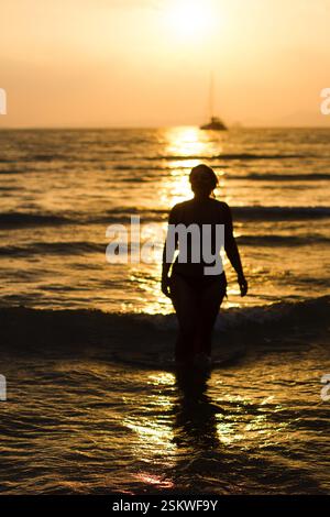 Silhouette einer Frau, die bei Sonnenuntergang im Meer mit einem Segelboot im Hintergrund und goldenen Reflexen auf dem Wasser am Railay Beach, Thailand, spaziert Stockfoto