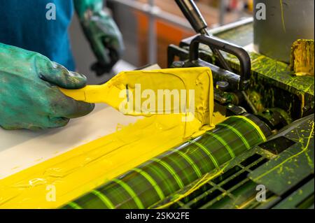 Druckerarbeiter verteilen während des Druckvorgangs gelbe Tinte mit einem Teigschaber auf einer industriellen CMYK-Druckermaschine Stockfoto
