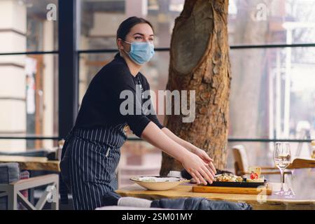 Kellnerin, die eine Gesichtsmaske trägt, bedient Kunden in einem Café Stockfoto