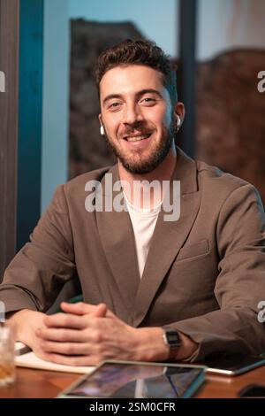 Ein Mann im Anzug lächelt und sitzt mit Laptop und Tablet an einem Tisch. Er arbeitet oder genießt seine Zeit am Tisch Stockfoto
