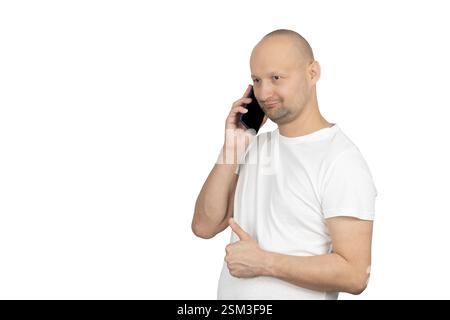 Ein kahlköpfiger Weißer mit kurzem Bart trägt ein weißes T-Shirt, hält ein Smartphone am Ohr, während er mit der anderen Hand die Daumen nach oben drückt. Er h Stockfoto