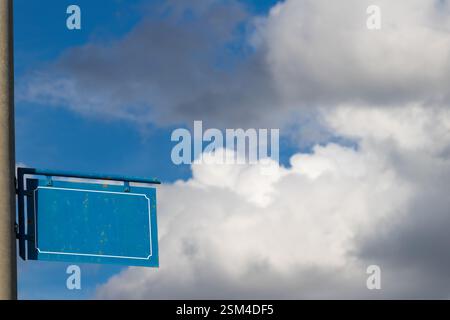 Blaues rechteckiges Metallschild mit dekorativer Borte auf einem Stab, vor einem Hintergrund mit flauschigen weißen Wolken und einem hellblauen Himmel, EV Stockfoto