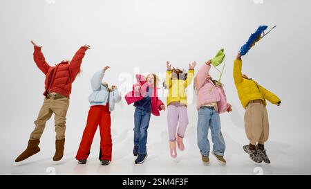 Gruppe von Kindern, die Arme angehoben, Hüte werfen, in helle Winterkleidung gekleidet, genießen energiegeladene Wintermomente vor weißem Studiohintergrund. Stockfoto