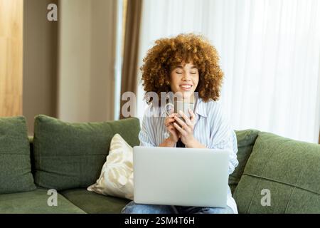 Eine junge Frau sitzt bequem auf einer weichen, grünen Couch und genießt einen Dampfbecher, während sie in einem sonnendurchfluteten Zuhause an ihrem Laptop arbeitet Stockfoto