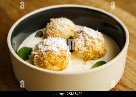Goldene, knusprig gebratene Hüttenkäse-Knödel, mit Puderzucker bestäubt, serviert in einer Schüssel mit cremiger Vanillesauce. Garniert mit frischen Minzblättern Stockfoto