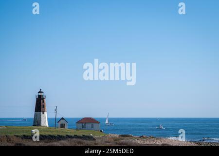 Ein malerischer Blick auf einen historischen Leuchtturm in der Nähe des Ozeans mit Booten und einem Segelboot in der Ferne unter einem klaren blauen Himmel an einem sonnigen Tag. Stockfoto