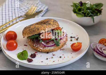 Leckerer Mohnbagel mit Räucherfleisch, cremigem Käse, Kirschtomaten, roten Zwiebeln und frischem Rucola auf einem weißen Teller mit Gewürzen Stockfoto