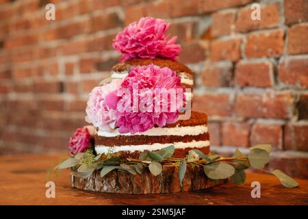 Ein atemberaubender dreistufiger Kuchen mit leuchtenden rosafarbenen Pfingstrosen sitzt auf einer Holzplatte vor einer Backsteinmauer. Stockfoto