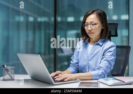 Ernsthafte, selbstbewusste asiatische Frau, die im Büro am Arbeitsplatz mit einem Laptop arbeitet. Frau tippt auf der Tastatur und bereitet einen elektronischen Bericht vor. Nachdenkliche und konzentrierte junge Geschäftsfrau, die am Tisch sitzt. Stockfoto