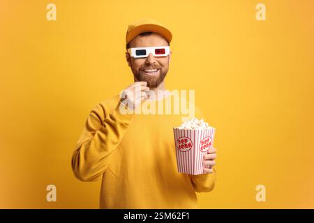 Glücklicher Mann in einem gelben Sweatshirt und einer Kappe mit 3D-Kino-Brille vor einem gelben Hintergrund, der eine Kiste Popcorn hält Stockfoto