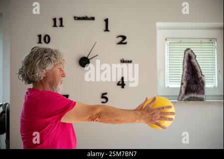 Ein älterer Mann mit lockigen grauen Haaren und einem Bart, der ein rosa Hemd trägt, dehnt und übt seine Arme nach vorne, während er einen gelben Pilates-Ball hineinhält Stockfoto