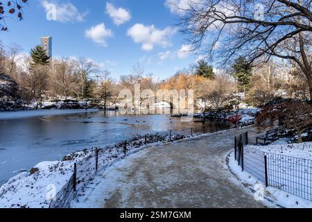 Der Schnee bedeckt den Boden, während der gefrorene Teich den Winterhimmel im Central Park neben der Gapstow Bridge reflektiert und so die ruhige Schönheit des Manhattan Winter noch verschönert Stockfoto