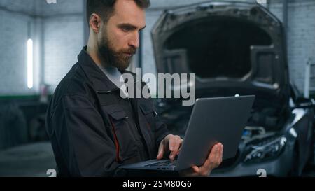 Ernsthaft fokussierter Kaukasier Guy Techniker Automechaniker Ingenieur prüfen Reparatur Autoproblem Computerdiagnose Elektromotor mit Stockfoto