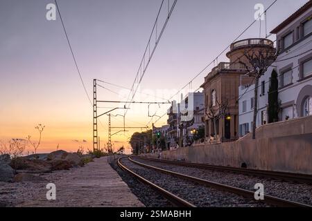 Eisenbahngleise entlang der Küste in Sant Pol de Mar, Katalonien, bei Sonnenuntergang, mit Gebäuden auf der rechten Seite, Felsen und Meer auf der linken Seite. Stockfoto