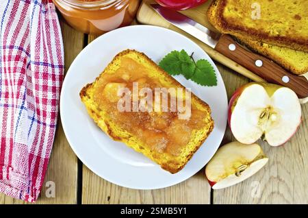 Scheibe Brot mit Apfelkonfitüre und Minze auf einem Teller, Apfelscheiben, ein Glas Marmelade, Serviette, Messer auf Holzbrett Hintergrund Stockfoto