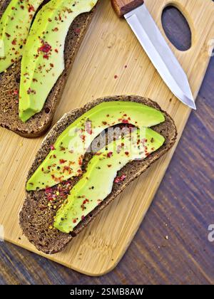 Zwei Scheiben Graubrot mit Scheiben von Avocado und Pfeffer, Messer gegen die dunkle Holzplatte Stockfoto