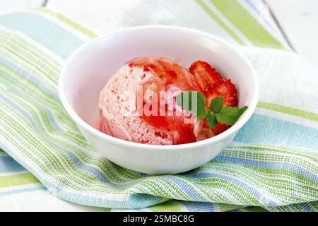 Erdbeereis in einer weißen Schüssel mit Erdbeeren und Erdbeersirup auf einer gestreiften Leinenserviette auf Holzbrettern Stockfoto