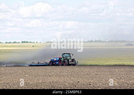 Blauer Radtraktor, der auf dem Hintergrund von gepflügtem Land, Getreide und Himmel arbeitet Stockfoto