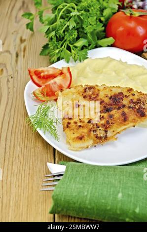 Gebratener Fisch mit Kartoffelbrei auf einem Teller, Petersilie, Tomaten, Serviette, Messer und Gabel auf einem Holzbrett Stockfoto